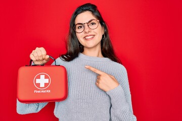 Young beautiful girl holding first aid kit medical box over red background very happy pointing with...
