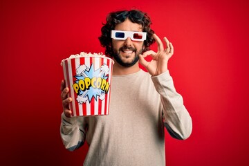 Young handsome man with beard watching movie holding popcorns over red background doing ok sign with fingers, excellent symbol