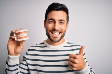 Young handsome man with beard holding plastic denture teeth over white background doing happy thumbs up gesture with hand. Approving expression looking at the camera showing success.