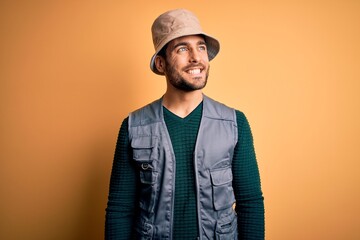 Handsome tourist man with beard on vacation wearing explorer hat over yellow background looking away to side with smile on face, natural expression. Laughing confident.