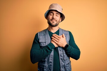 Handsome tourist man with beard on vacation wearing explorer hat over yellow background smiling with hands on chest with closed eyes and grateful gesture on face. Health concept.