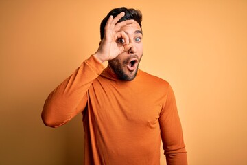 Young handsome man with beard wearing casual sweater standing over yellow background doing ok gesture shocked with surprised face, eye looking through fingers. Unbelieving expression.