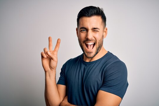Young handsome man wearing casual t-shirt standing over isolated white background smiling with happy face winking at the camera doing victory sign. Number two.