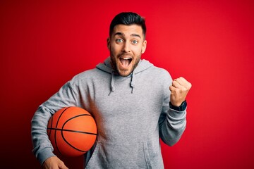 Young sports man holding basketball ball over red isolated background screaming proud and...