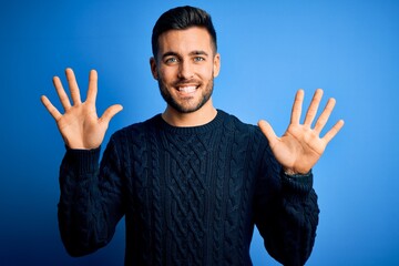 Young handsome man wearing casual sweater standing over isolated blue background showing and pointing up with fingers number ten while smiling confident and happy.