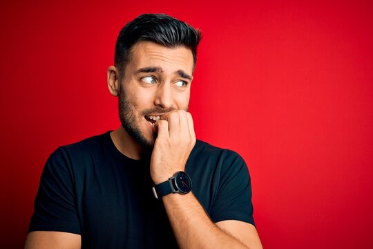 Young handsome man wearing casual black t-shirt standing over isolated red background looking stressed and nervous with hands on mouth biting nails. Anxiety problem.