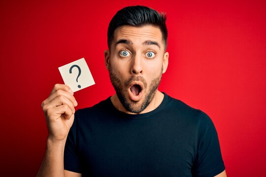 Young handsome man holding paper with question mark symbol over red background scared in shock with a surprise face, afraid and excited with fear expression