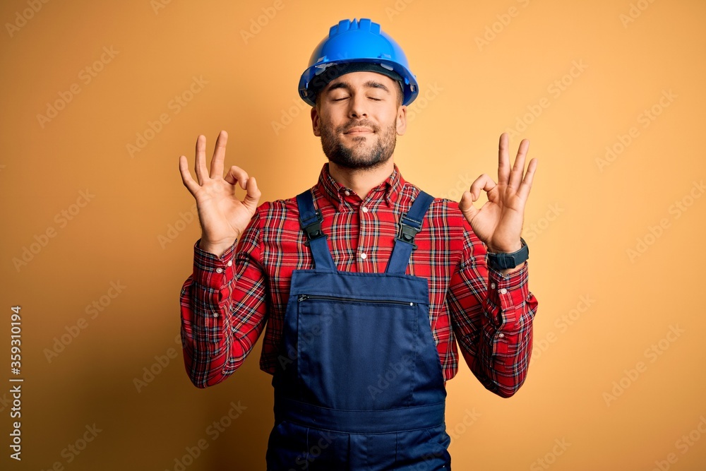 Wall mural young builder man wearing construction uniform and safety helmet over yellow background relax and sm