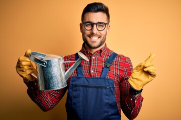 Young gardener man wearing working apron and gloves holding watering can very happy pointing with hand and finger to the side © Krakenimages.com