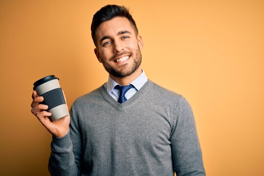 Young handsome man drinking a hot coffee from take away plastic cup over yellow background with a happy face standing and smiling with a confident smile showing teeth