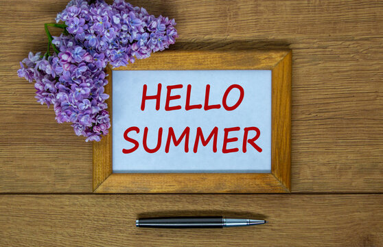 Wooden picture frame with inscription 'hello summer' on beautiful wooden table, lilac branch and metalic pen. Concept.