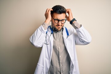 Young doctor man wearing glasses, medical white robe and stethoscope over isolated background suffering from headache desperate and stressed because pain and migraine. Hands on head.