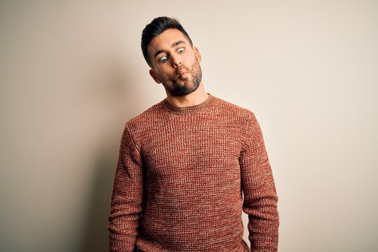 Young handsome man wearing casual sweater standing over isolated white background making fish face with lips, crazy and comical gesture. Funny expression.