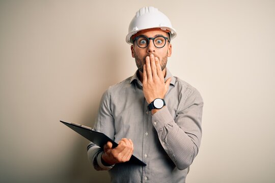 Young architect inspector man wearing builder safety helmet and using clipboard cover mouth with hand shocked with shame for mistake, expression of fear, scared in silence, secret concept