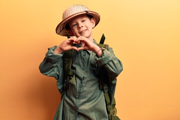 Cute blond kid wearing explorer hat and backpack smiling in love doing heart symbol shape with hands. romantic concept.