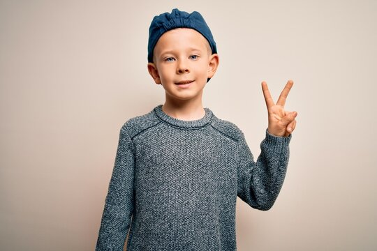 Young little caucasian kid with blue eyes wearing wool cap over isolated background smiling with happy face winking at the camera doing victory sign. Number two.