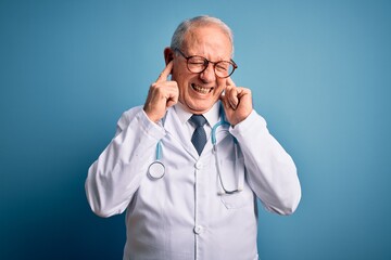 Senior grey haired doctor man wearing stethoscope and medical coat over blue background covering ears with fingers with annoyed expression for the noise of loud music. Deaf concept.