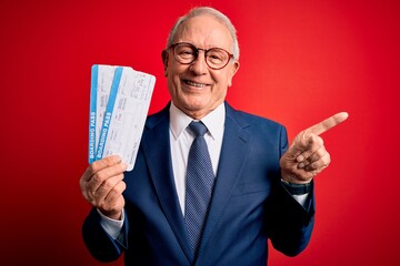 Senior grey haired business man holding airplane boarding pass over red background very happy pointing with hand and finger to the side
