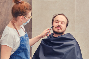 A woman in medical mask shaves a man beard with a safety razor