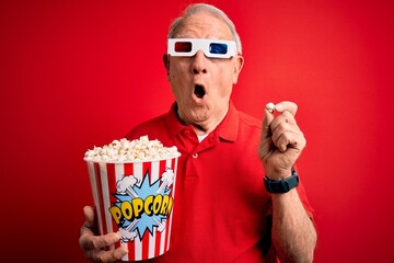Grey haired senior man wearing 3d movie glasses and eating popcorn over red background scared in...