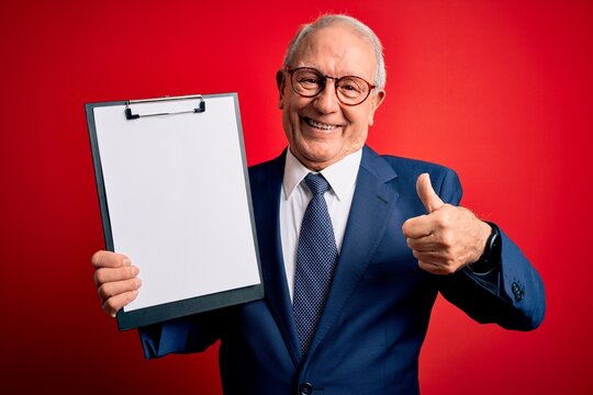 Senior grey haired business man holding clipboard over red background happy with big smile doing ok sign, thumb up with fingers, excellent sign