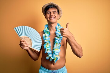 Young tourist man on vacation wearing swimwear and hawaiian lei flowers holding hand fan happy with big smile doing ok sign, thumb up with fingers, excellent sign