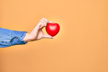 Hand of caucasian young woman holding red heart over isolated yellow background