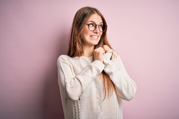 Young beautiful redhead woman wearing casual sweater and glasses over pink background laughing nervous and excited with hands on chin looking to the side