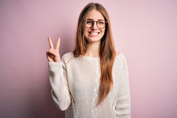 Young beautiful redhead woman wearing casual sweater and glasses over pink background showing and pointing up with fingers number two while smiling confident and happy.
