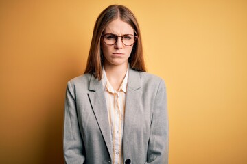 Young beautiful redhead woman wearing jacket and glasses over isolated yellow background skeptic...
