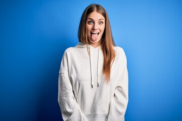 Young beautiful redhead sporty woman wearing sweatshirt over isolated blue background sticking tongue out happy with funny expression. Emotion concept.