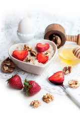 Breakfast with oatmeal with strawberries, honey, walnuts and egg on white background. Breakfast for child. Oatmeal in a white heart-shaped bowl