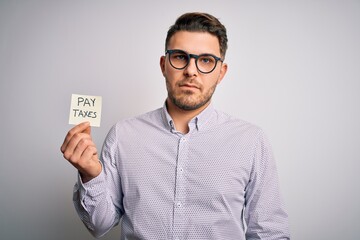 Young business man with blue eyes holding pay taxes word on paper note with a confident expression...