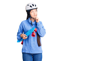 Young beautiful chinese woman wearing security helmet holding skate serious face thinking about question with hand on chin, thoughtful about confusing idea