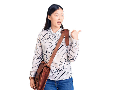 Young beautiful chinese woman holding briefcase pointing thumb up to the side smiling happy with open mouth