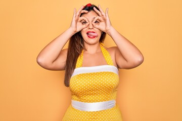 Young hispanic pin up woman wearing fashion sexy 50s style over yellow background doing ok gesture like binoculars sticking tongue out, eyes looking through fingers. Crazy expression.