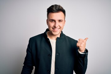 Young handsome business mas wearing elegant winter coat standing over isolated background smiling with happy face looking and pointing to the side with thumb up.