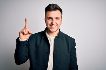 Young handsome business mas wearing elegant winter coat standing over isolated background showing and pointing up with finger number one while smiling confident and happy.