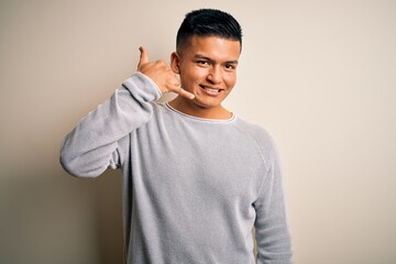 Young handsome latin man wearing casual sweater standing over isolated white background smiling doing phone gesture with hand and fingers like talking on the telephone. Communicating concepts.
