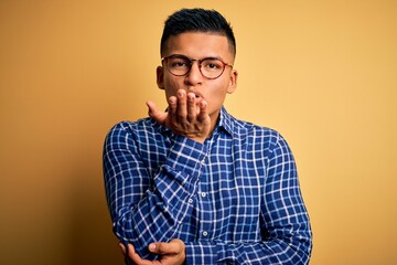 Young handsome latin man wearing casual shirt and glasses over yellow background looking at the camera blowing a kiss with hand on air being lovely and sexy. Love expression.