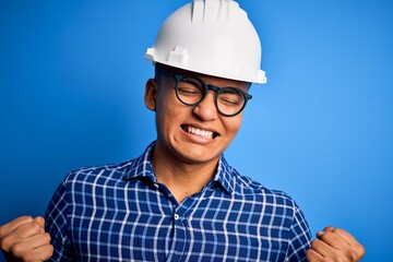 Young handsome engineer latin man wearing safety helmet over isolated blue background very happy and excited doing winner gesture with arms raised, smiling and screaming for success. Celebration