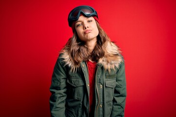 Young blonde girl wearing ski glasses and winter coat for ski weather over red background looking at the camera blowing a kiss on air being lovely and sexy. Love expression.