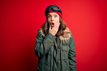 Young blonde girl wearing ski glasses and winter coat for ski weather over red background Looking fascinated with disbelief, surprise and amazed expression with hands on chin