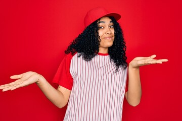 Young african american curly sportswoman wearing baseball cap and striped t-shirt clueless and confused expression with arms and hands raised. Doubt concept.