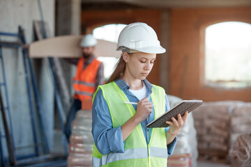 A construction industry maintenance engineer is a pretty woman dressed in a uniform and protective helmet with a tablet in her hands against the background of a construction site and a worker