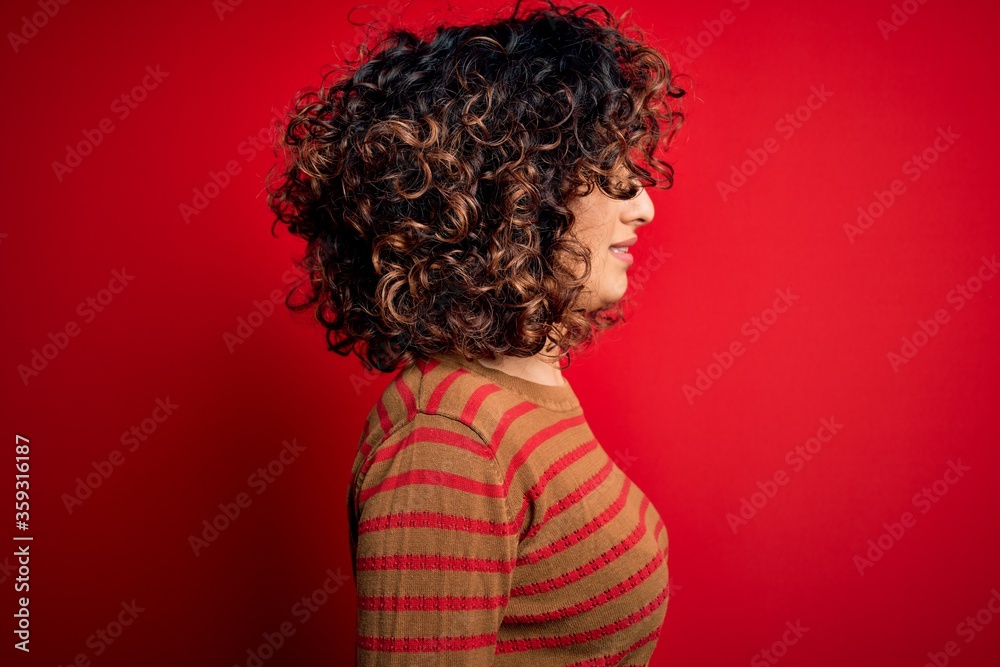 Sticker young beautiful curly arab woman wearing casual striped sweater standing over red background looking