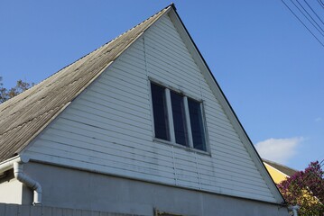 white wooden attic of a rural house with one window under a gray slate roof against a blue sky