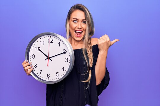 Young beautiful blonde woman doing countdown using big clock over purple background pointing thumb up to the side smiling happy with open mouth