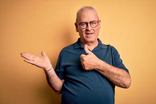 Middle age handsome hoary man wearing casual polo and glasses over yellow background Showing palm hand and doing ok gesture with thumbs up, smiling happy and cheerful