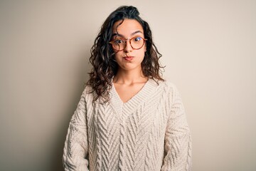 Beautiful woman with curly hair wearing casual sweater and glasses over white background puffing cheeks with funny face. Mouth inflated with air, crazy expression.